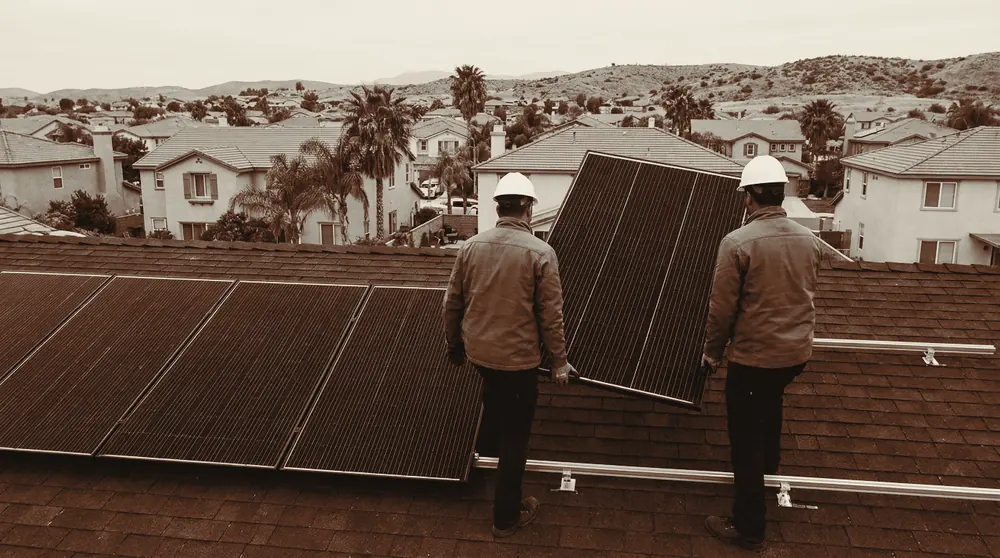 Two workers in hard hats lower a solar panel onto an aluminium mounting rail on a shingle roof, beside a row of panels already fixed in place.